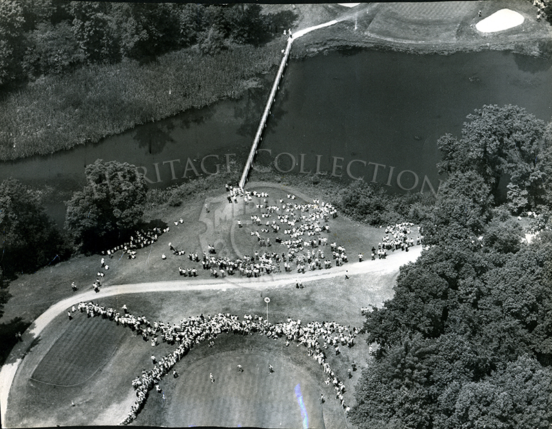 Aerial view from June 11, 1949, pictures a scene from the 49th United States Open at Medinah Country Club. The image shows the gallery watching two foursomes on 16th hole (below) and the 17th greens. The tee shot is across a wind-swept Lake Kadijah and the green is surrounded by sand traps and woods.