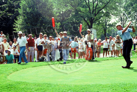Charles Coody is photographed in full swing during the Ninth U.S. Senior Open Championship. On the left, Chi Chi Rodriguez, wearing white hat, white shirt and blue pants, heads toward the fairway. The sign in the background shows that Coody and Rodriguez were grouped with Lou Graham. Note the Ã´Quite PleaseÃ¶ paddles held up by two of the 276 volunteers that served during the tournament.